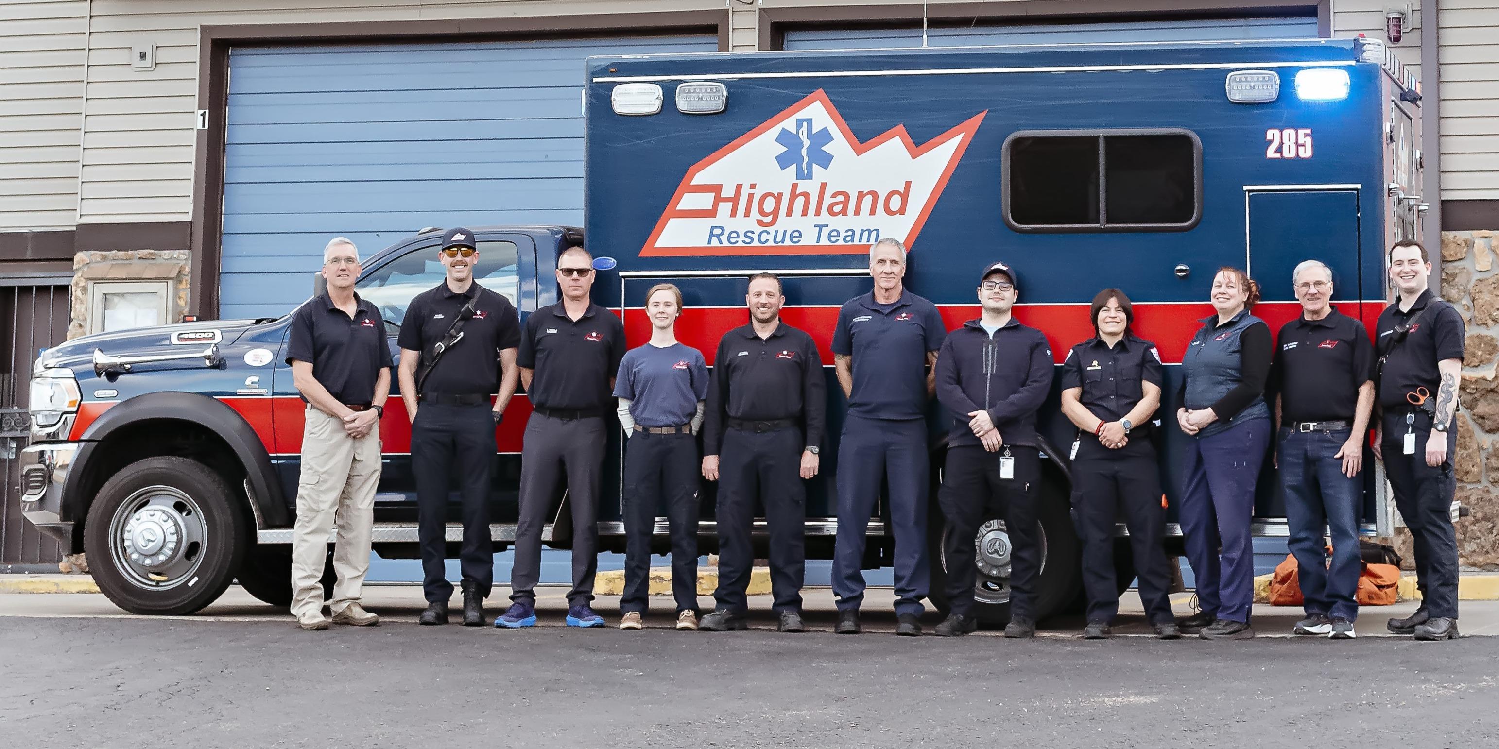 a group of EMS professionals in navy blue uniforms stand proudly in front of a navy blue ambulance with a red stripe